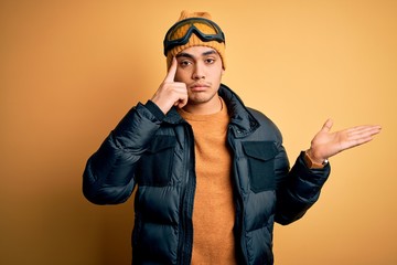 Young brazilian skier man wearing snow sportswear and ski goggles over yellow background confused and annoyed with open palm showing copy space and pointing finger to forehead. Think about it.