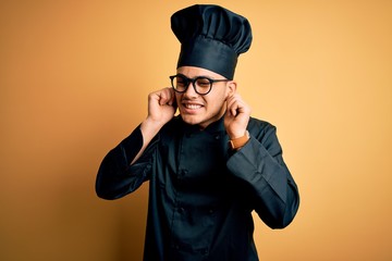 Young brazilian chef man wearing cooker uniform and hat over isolated yellow background Smiling pulling ears with fingers, funny gesture. Audition problem
