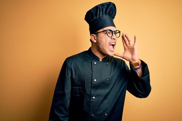 Young brazilian chef man wearing cooker uniform and hat over isolated yellow background shouting and screaming loud to side with hand on mouth. Communication concept.