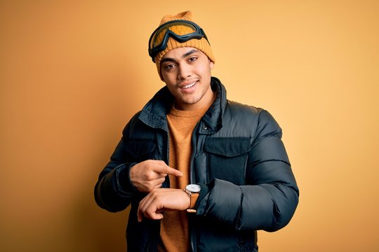 Young Brazilian Skier Man Wearing Snow Sportswear And Ski Goggles Over Yellow Background In Hurry Pointing To Watch Time, Impatience, Upset And Angry For Deadline Delay