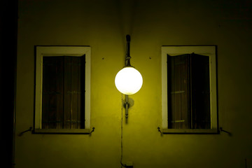Lampada illuminazione pubblica in centro storico della città illumina un muro con due finestre chiuse con balconi in legno.