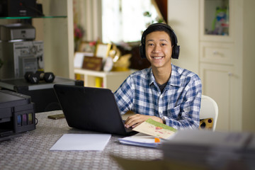 Happy young Asian teenage of a boy wear wireless headset studying online class on laptop computer at home smiling to camera. - Powered by Adobe