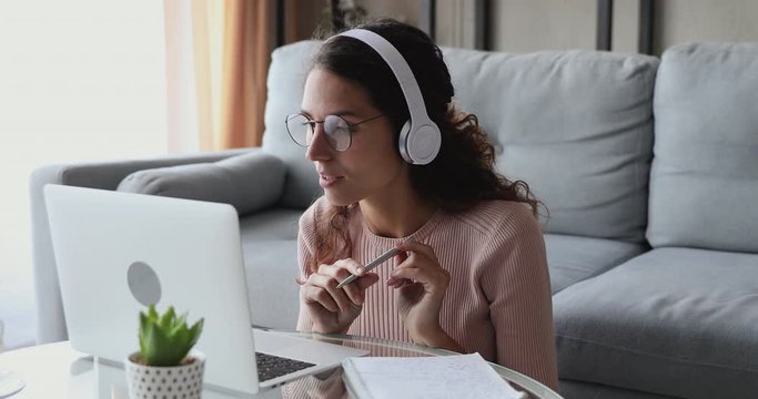 Young woman wearing headphones distance learning course, doing remote telework concept working from home office. Female student, online teacher conference calling by web cam using laptop making notes.