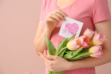 Woman holding greeting card and flowers for Mother's Day on color background