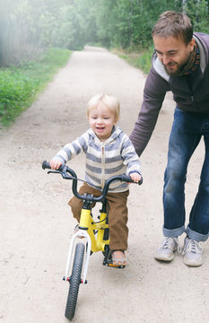 Cute smiling blond boy learning to ride bicycle. Father teaching his little child to ride bike in spring summer park. Happy family moments. Time together dad and son. Candid lifestyle image.