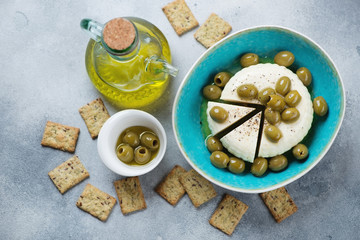 Fototapeta na wymiar Soft cheese with green olives, olive oil and crackers. Above view on a light-blue stone background, horizontal shot