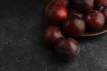 Blue plums scattered from ceramic brown plate on a dark stone table. Blue ripe plums on textured dark grey background. Copy space. 