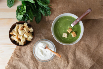 Healthy green soup puree in a grey bowl with crouton, raw spinach leaves and sour cream with spoon on brown sackcloth on wooden board. Top view