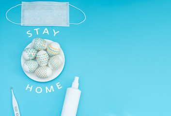 Decorated Easter eggs lie on the plate, medical face mask, thermometer and sanitizer on blue background with copy space. Happy Easter during quarantine, stay home, social distance, COVID-19 concepts.