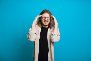 Caucasian man with long hair and bristle is gesturing a headache touching his head on a blue wall