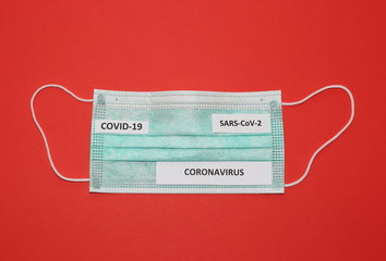 Surgical protective face mask with name of illness CORONAVIRUS, covid-19 and SARS-CoV-2 on it top view close up on red background