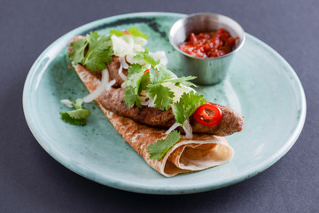 Meat sausages with fresh greenery on lavash bread with sauce in a sauce pan served on a blue plate over dark background.