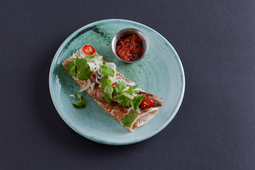 Meat sausages with fresh greenery on lavash bread with sauce in a sauce pan served on a blue plate over dark background.