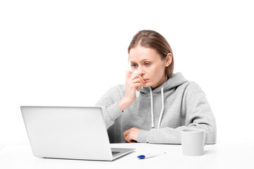 Sick young woman with blowing nose sitting by desk in front of laptop