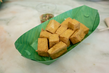 Deep fried tofu is cut into square balls in a green paper dish. The side has a cup of sauce. Is a Chinese and Japanese style food.