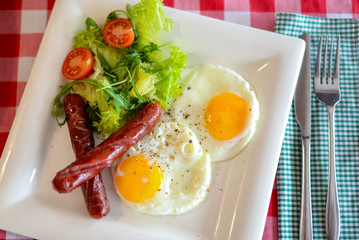 Breakfast with salad, fried egg and sausages served in white plate over red plate tablecloth.