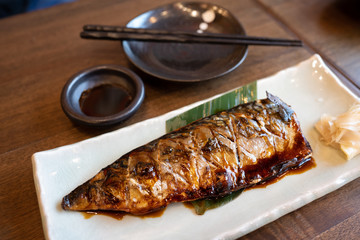 Grilled saba fish with teriyaki sauce on a white plate on a wooden table with a bowl of sauces and chopsticks Is a popular Japanese food.