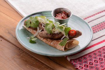 Meat sausages with fresh greenery on lavash bread with sauce in a sauce pan served on a blue plate over wooden table.