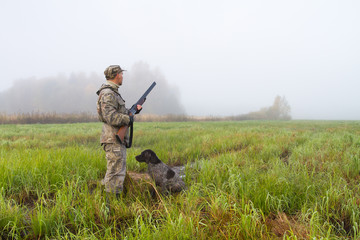 a hunter and his assistant dog are in a meadow on a foggy morning