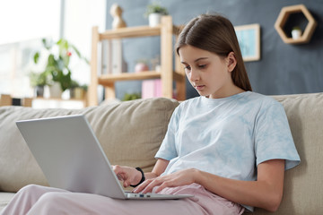 Teenage girl in casualwear sitting on couch with laptop on her knees at home