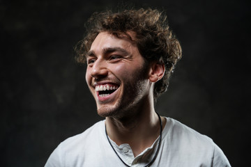 Fototapeta premium Portrait of a smiling young adult guy with curly haircut, wearing casual white shirt and necklace. Looking on the side while laughing