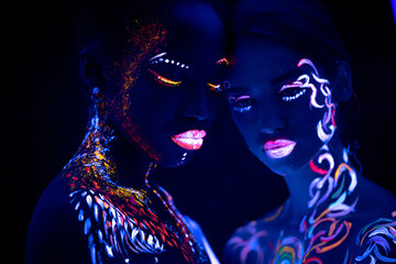 young beautiful girls with fluorescent make-up posing at camera, unusual creative prints on their skin. attractive women hug each other. prints glows in UV light.isolated black background