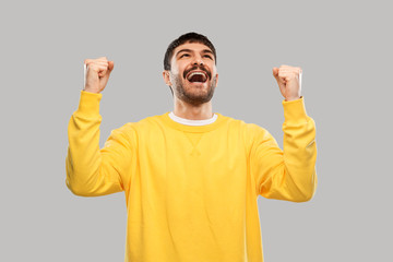 success, emotion and expression concept - happy man in yellow sweatshirt celebrating victory over grey background