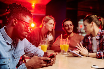 African guy scrolling in smartphone while sitting by desk in cafe with friends