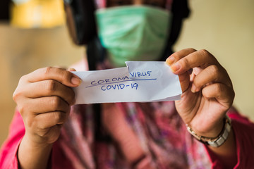 Woman wearing a surgical mask rips off a small piece of paper with a handwritten note that reads coronavirus and covid-19