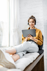 Young woman with a headset working online on a digital tablet while sitting on the window sill at...