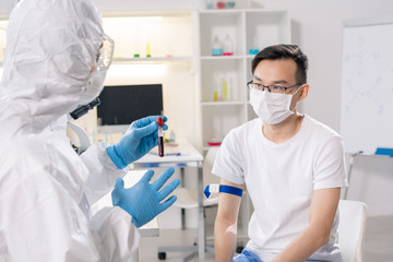 Sick chinese guy in protective mask sitting in front of lab worker with flask