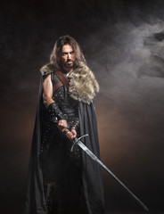 Man dressed in medieval armour and raincoat with longs word over smoke background. Courage fantasy warrior knight with long hair concept historical photo