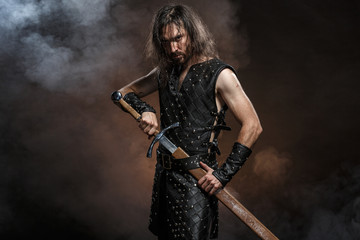 Man dressed in medieval armour and raincoat with longs word over smoke background. Courage fantasy...