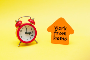 Red alarm clock house shaped orange sticky note on yellow background. Work from home concept. Social distancing, pandemic, corona virus, home office time, self-isolation. Digital nomads.