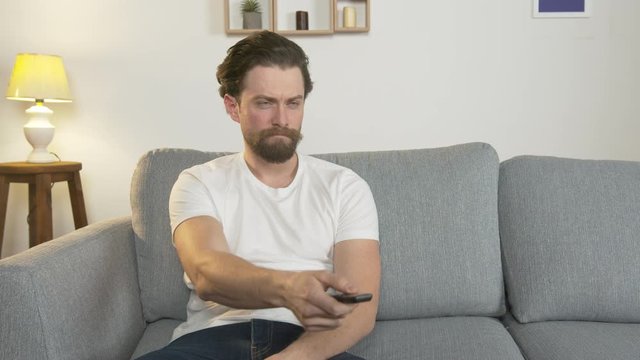 bored man sits alone on a sofa in a room and watches TV, holds a remote control in his hand, switches channels, isolation.
