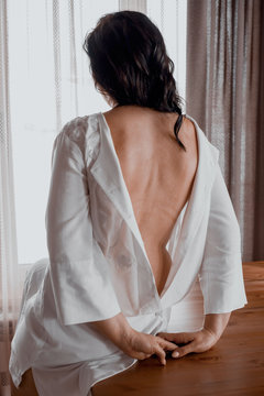 A woman is sitting on a table in a white tunic. Rear view
