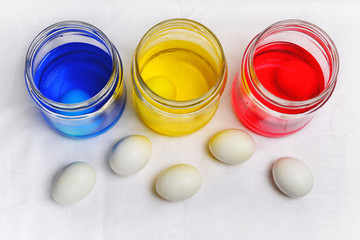 Three glass cans with blue yellow and red colorings for easter eggs proceeding and five white chicken eggs scattered on a linen tablecloth