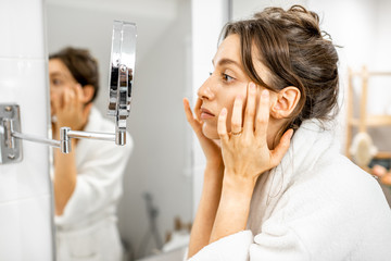 Young woman in bathrobe taking care of her skin, looking at the mirror and worrying about wrinkles...