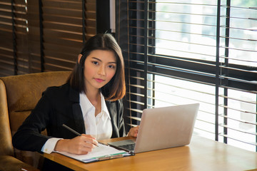 portrait young charming Asian woman in formal suit sitting in front of laptop and looking a camera, she is holding a pen for note something on check page on wooden table next a window