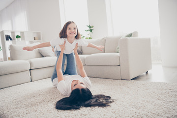 Photo of pretty funny little girl young mommy holding daughter hands above floor having fun pretending airplane flight lying carpet spend time weekend together home indoors