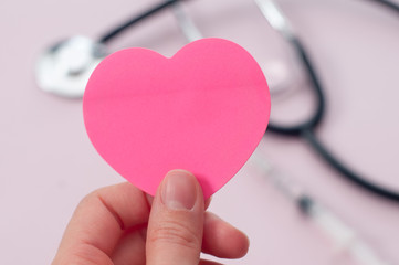 Woman hand holding heart shaped sticky note on medical equipment. Blurred stethoscope and syringe...