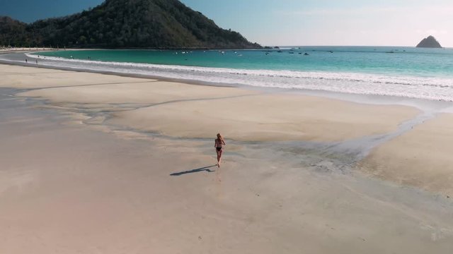 Aerial 4K drone shot during daylight of a young woman running at the beach into the water at the tropical remote surf beach Pentai Selong Belanak in Lombok, West-Nusa Tenggara, Indonesia