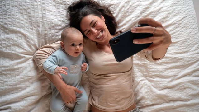 Authentic close up of neo mother and her newborn baby making a selfie or video call to father or relatives in a bed. Concept of technology, new generation,family, connection, parenthood, authenticity