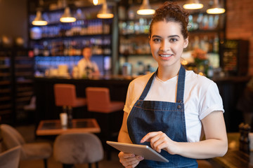 Young smiling waitress in apron and t-shirt standing in front of camera - obrazy, fototapety, plakaty
