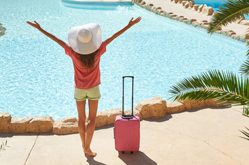 Travel, summer holidays and vacation concept - Beautiful woman raises her hands up near hotel pool area with pink suitcase