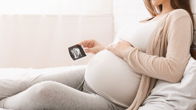 Pregnant woman holding ultrasound picture near belly, closeup