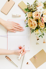 Vertical picture. Female hands wtite in pink opened notebook on white background. Tender mood and blogging concept with beautiful roses bouquet and accesoires in flat lay style