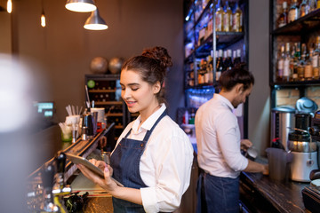 Pretty young waitress with tablet scrolling through online orders by workplace