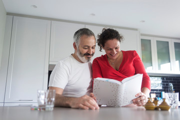 Obraz premium Happy pregnant woman with her husband sitting at home. Smiling couple consulting a book at the kitchen.