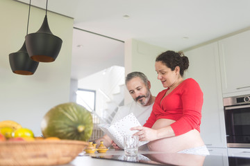 Happy pregnant woman with her husband sitting at home. Smiling couple consulting a book at the kitchen.